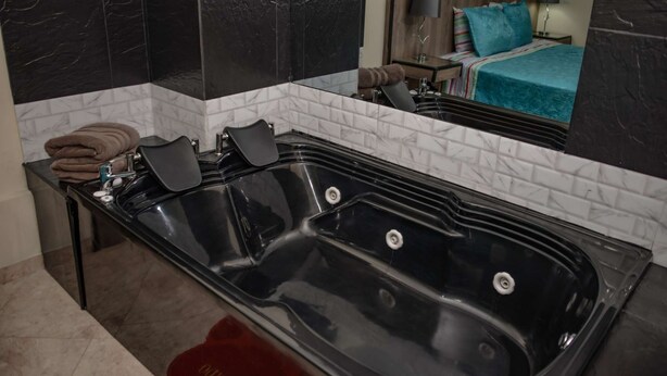 A modern bathroom with a large black bathtub, white marble tiles, and a mirror reflecting a bedroom with a bed and nightstand. - King One bedroom AC Hot tub elevator (Medellin)