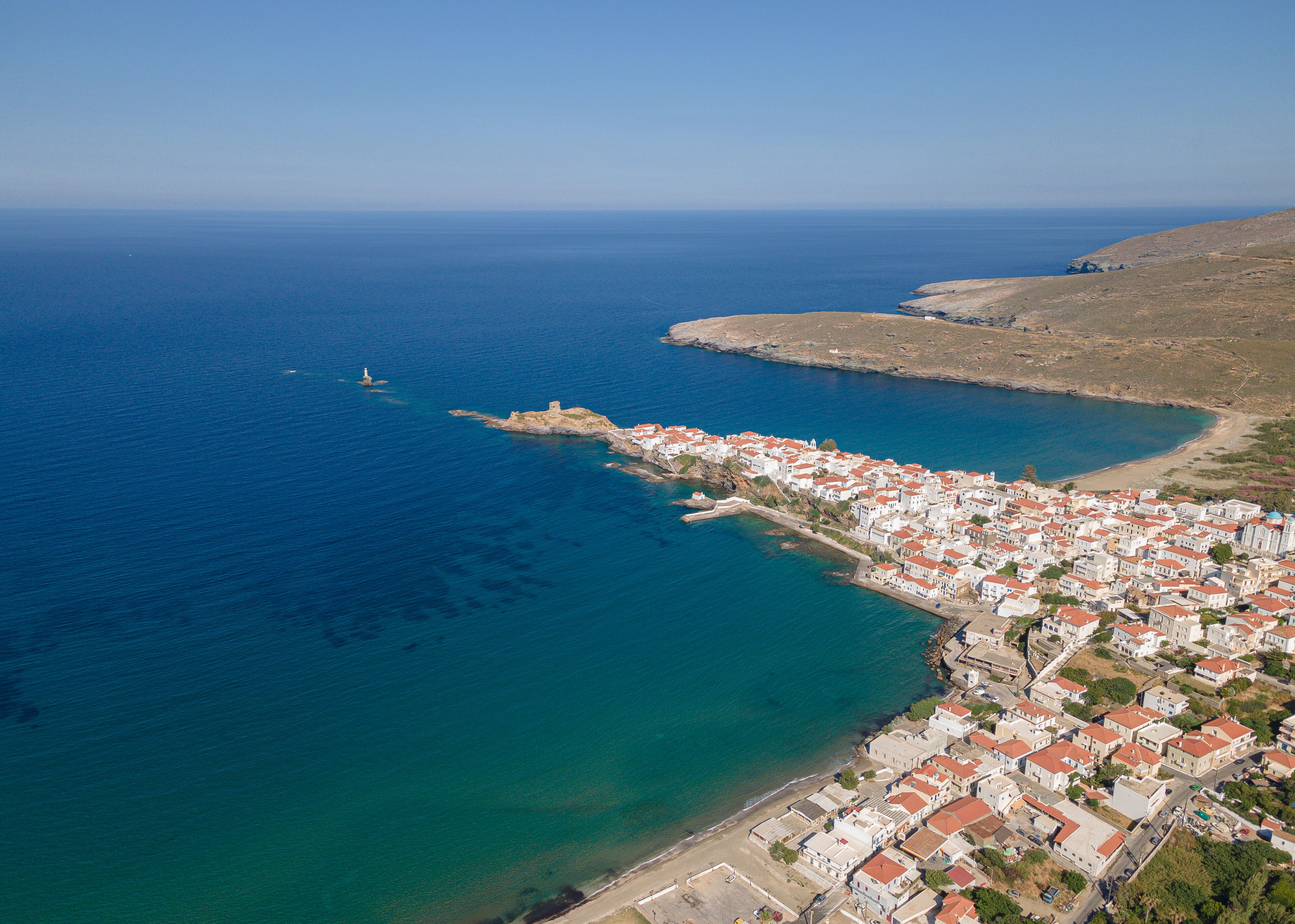 Une vue aérienne d’une ville côtière, avec un ensemble dense de bâtiments le long de la côte, un phare à l’horizon et une mer bleue et limpide.
