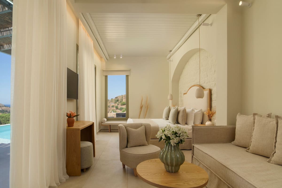 A modern hotel room with a bed, sofa, and armchair, a wooden side table, and a view of the ocean.