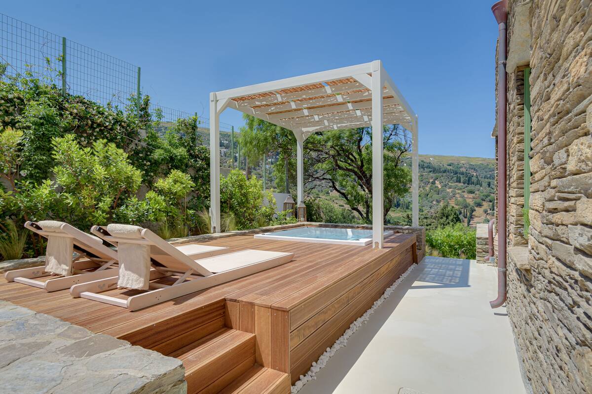 A wooden deck with lounge chairs and a pergola, a swimming pool, and a stone wall.