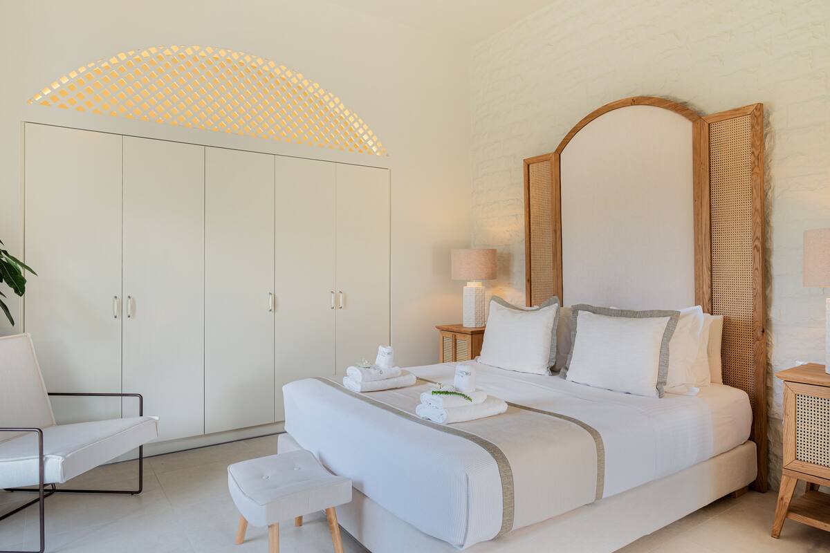 A modern bedroom with a large bed, a white wardrobe, a small desk, and a potted plant.