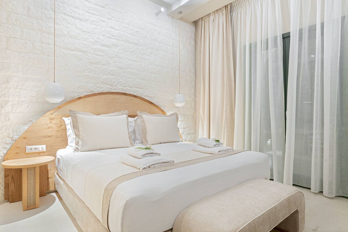 A modern bedroom with a large bed, a wooden headboard, a bedside table, and a large window with curtains.