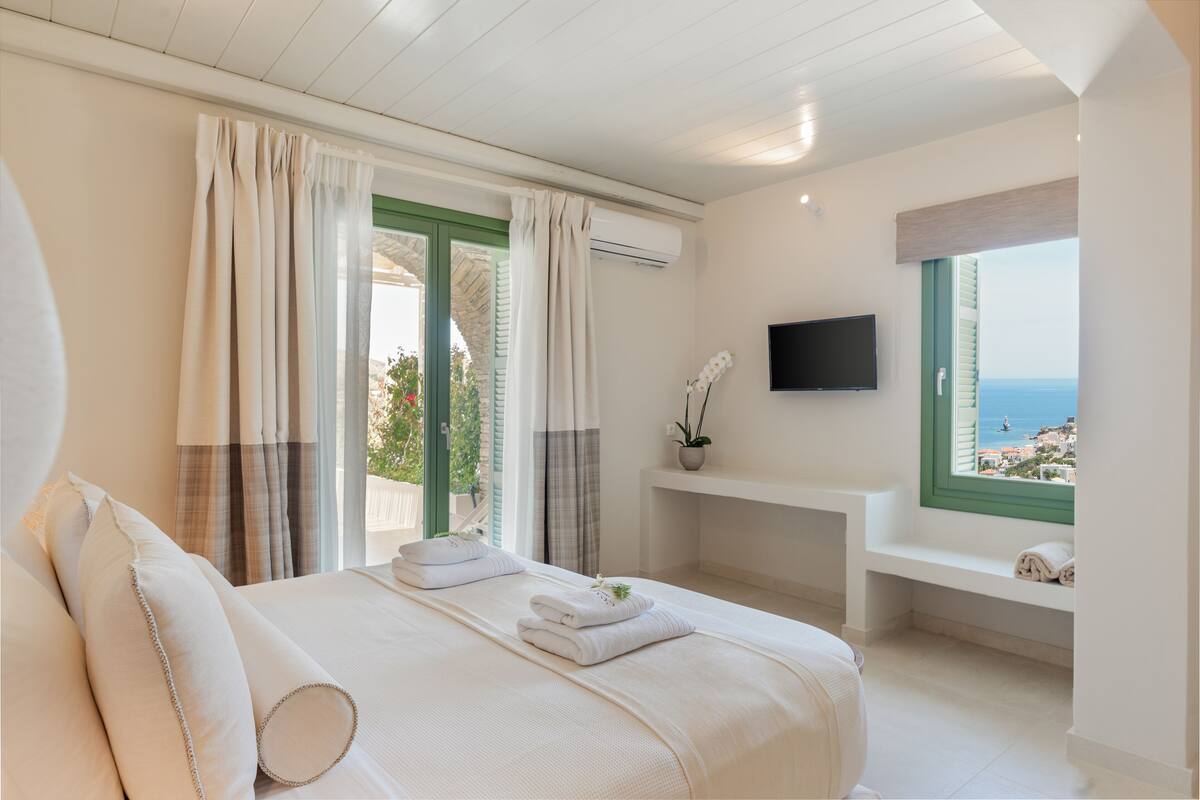 A bedroom with a bed, curtains, a TV, a window with a view, and a bench.