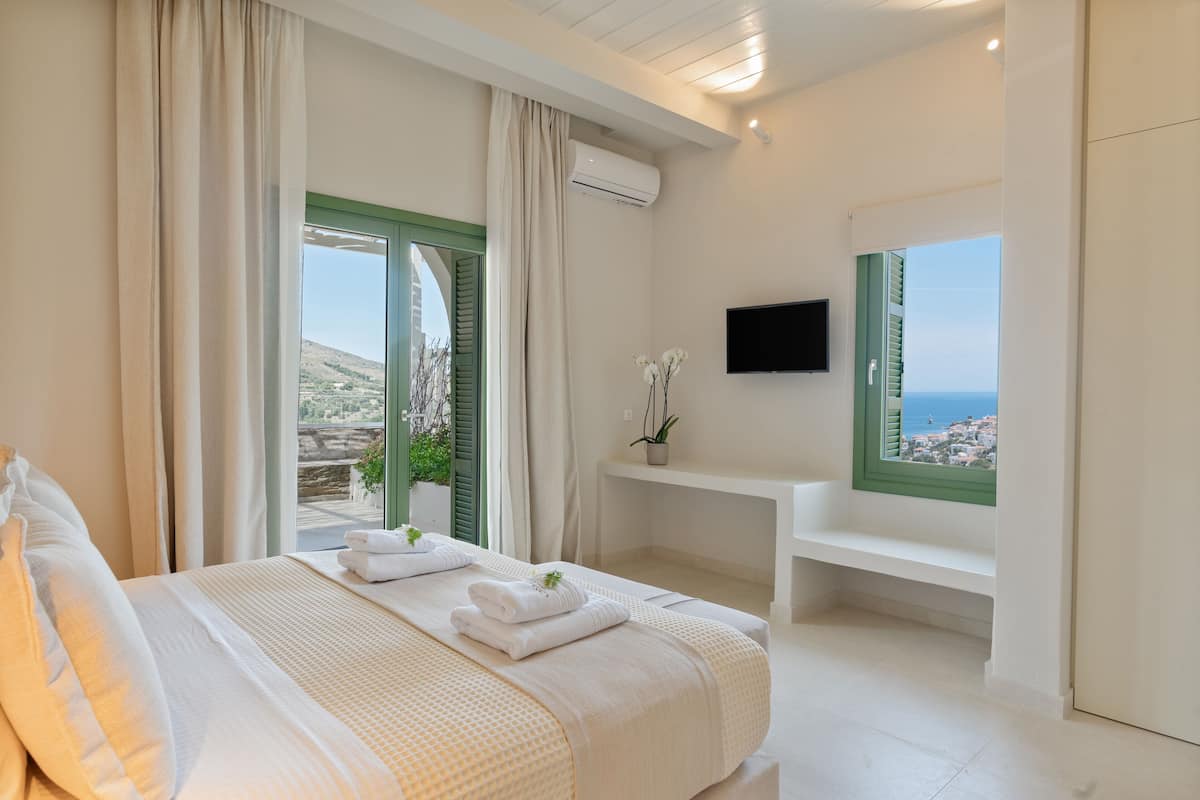 A bedroom with a bed, a TV, a window with a view of the sea, and a balcony.