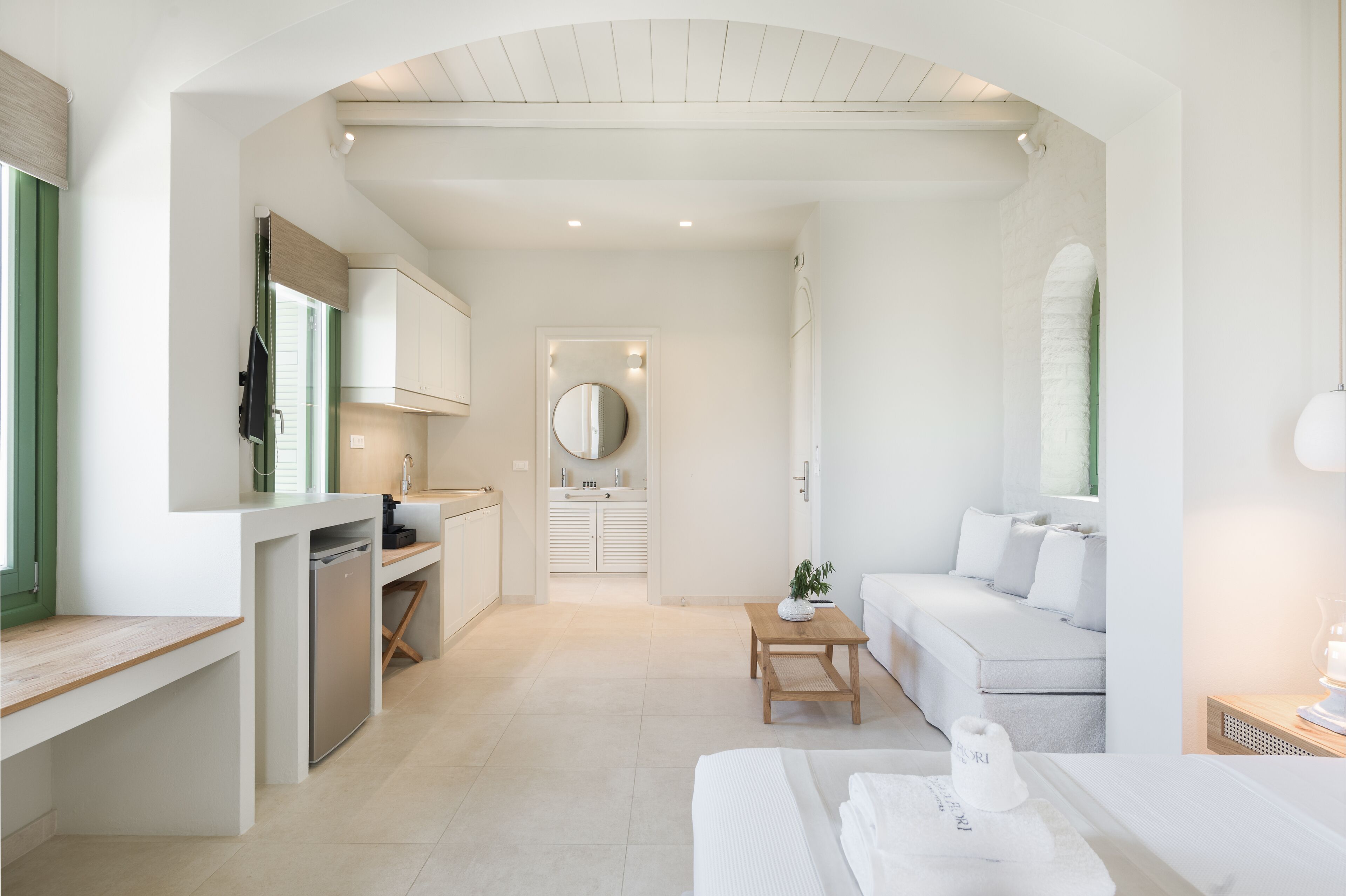A modern, compact hotel room with a kitchenette, a small dining area, a sofa, and a bathroom with a mirror and sink.