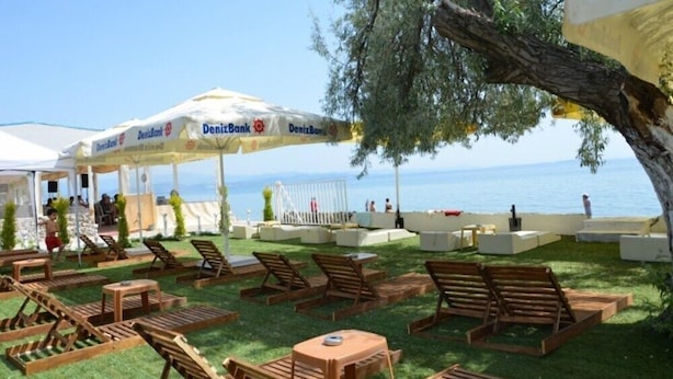 An outdoor seating area with wooden loungers and tables, a covered area with umbrellas, and a view of the sea. - Sir Motel (Edremit)