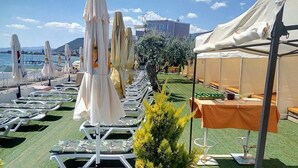 A seaside terrace with rows of sun loungers and umbrellas, a small table with bottles, and a view of the sea and buildings in the background. - Sir Motel (Edremit)