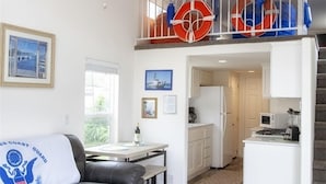 Un salón espacioso con ventilador de techo, un sofá de cuero negro, una mesa de centro y una zona de cocina con un refrigerador blanco y microondas.
