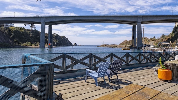 Una terraza de madera con sillas y una planta en maceta, con vista a un cuerpo de agua y un puente al fondo.