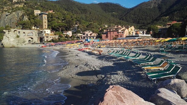 Ein Strand mit Reihen von Liegestühlen und Sonnenschirmen, felsiges Ufer und Gebäude im Hintergrund.