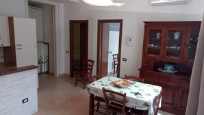 Ein Essbereich mit einem Tisch, gedeckt für vier Personen, umgeben von Holzstühlen. Rechts befindet sich ein Schrank mit Glastüren, links ein weißer Kühlschrank.