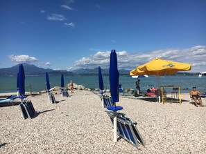 A pebble beach with blue and yellow umbrellas, lounge chairs, and a clear view of the sea and mountains in the background.