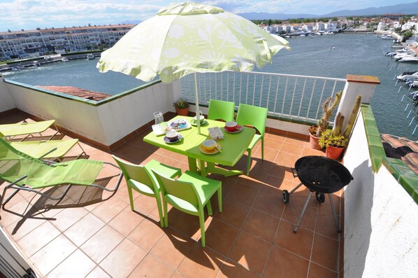 A terrace with a table, chairs, a grill, and a view of a marina with boats.