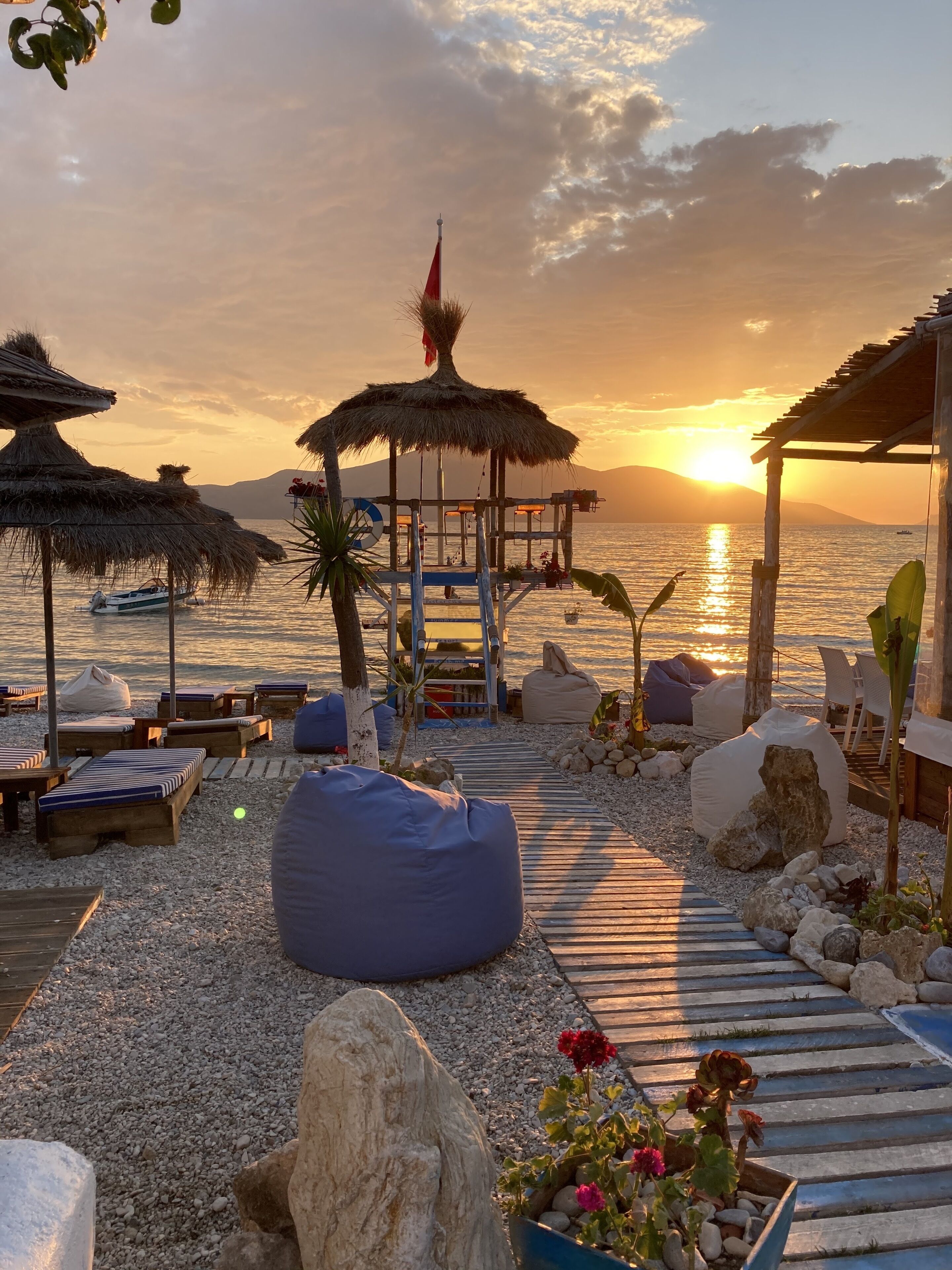 Un'area relax in spiaggia con ombrelloni di paglia, poltrone lounge e un pontile in legno che conduce a un molo.
