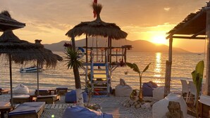 Un espace de détente aménagé sur la plage, avec des parasols en chaume, des chaises longues et une terrasse en bois menant à une jetée.