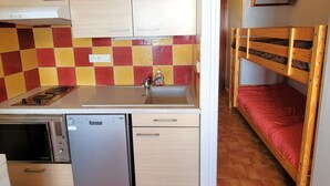A compact kitchen with a dishwasher, a microwave, and a sink. There is a bunk bed with a red mattress and wooden headboard.