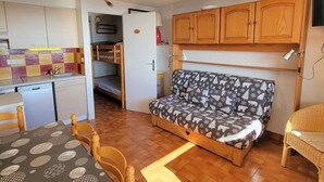 A compact room with a kitchenette, a bed with a patterned cover, wooden cabinets, and a dining area with a table and chairs.