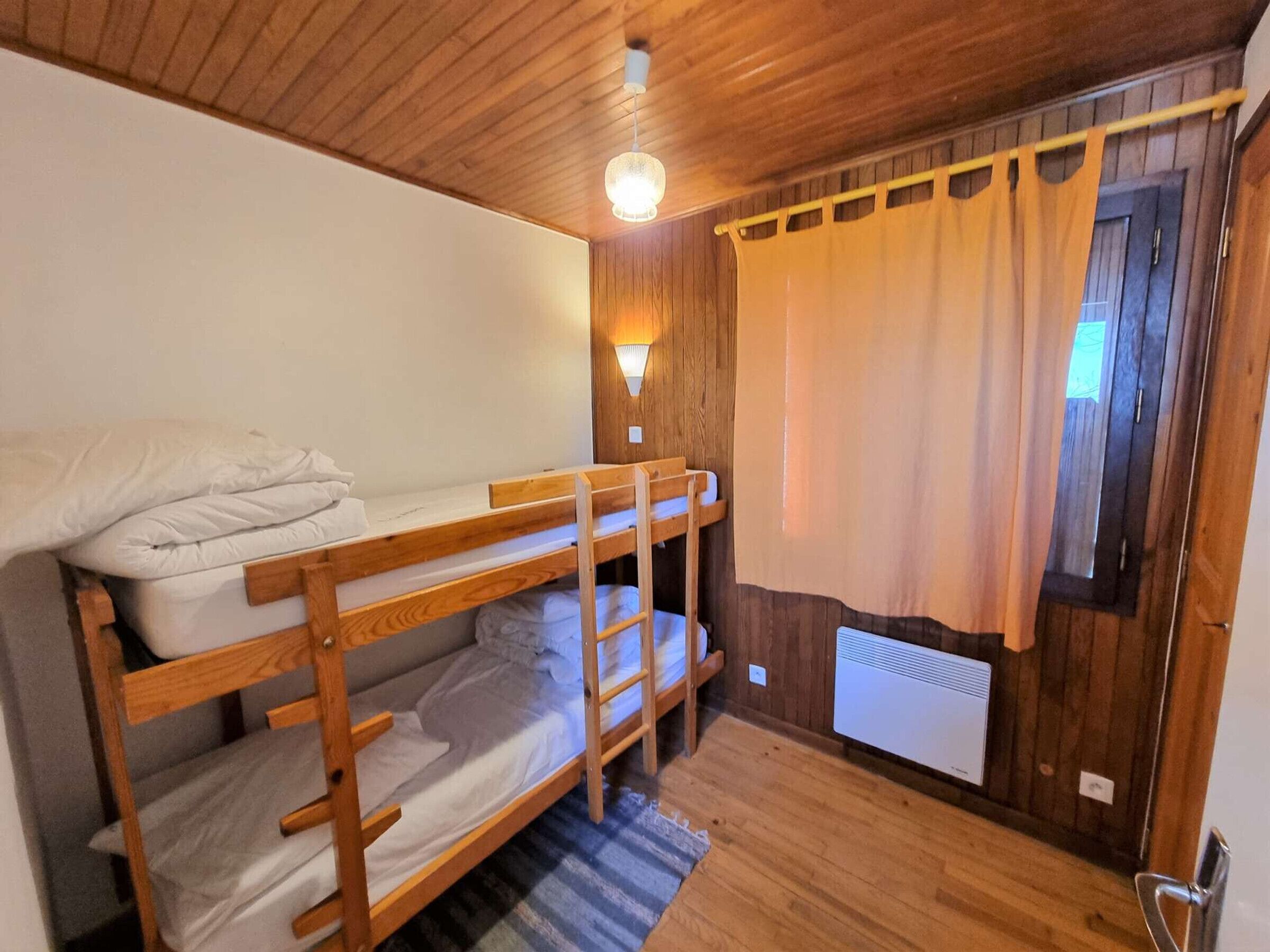 Dormitorio con literas, paredes de madera, una lámpara en el techo, un radiador y una cortina.