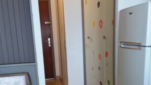 Un couloir avec un réfrigérateur, une porte ornée d’autocollants colorés et une fenêtre avec des stores.