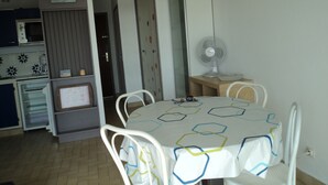 Une cuisine compacte équipée d’un four à micro-ondes, d’un petit réfrigérateur et d’une machine à café. On y trouve une table à manger avec des chaises et une nappe blanche. Un ventilateur blanc est posé sur une table en bois, à côté de l’espace repas.
