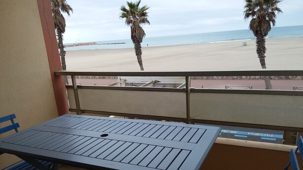 Un balcon avec une table et des chaises, donnant sur une plage bordée de palmiers et sur la mer.