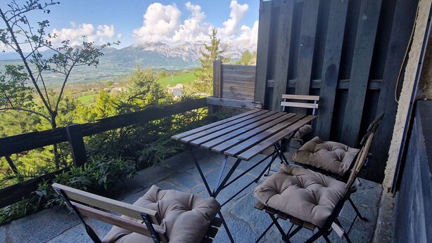 A patio with a wooden table and two cushioned chairs, overlooking a mountainous landscape.