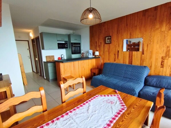 A living room with a wooden dining table, blue sofa, and a kitchen area with a microwave and oven.