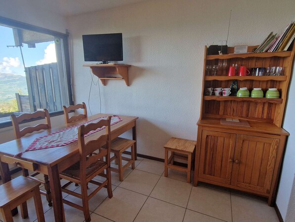 Un comedor con una mesa y sillas de madera, un televisor montado en la pared y un armario con cristalería y vajilla.