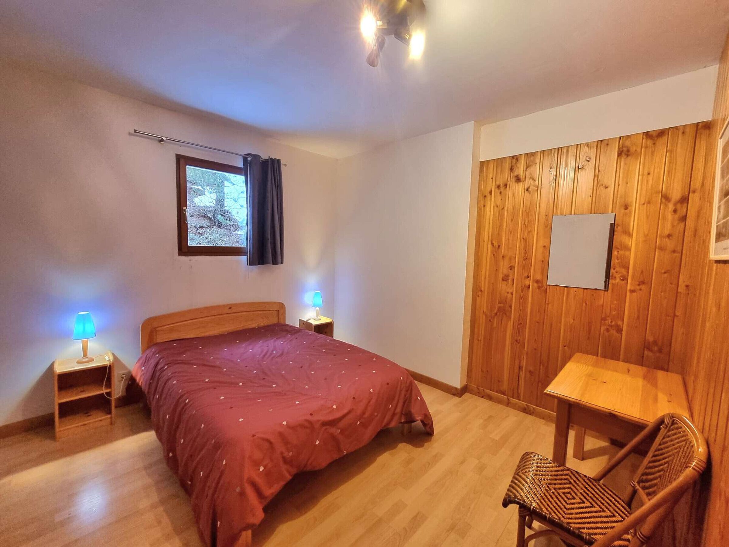 Een slaapkamer met een houten bed, bedlampjes, een klein tafeltje en een stoel.