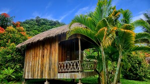 Una cabaña de madera con techo de paja, un porche de madera y un exuberante jardín verde con palmeras.