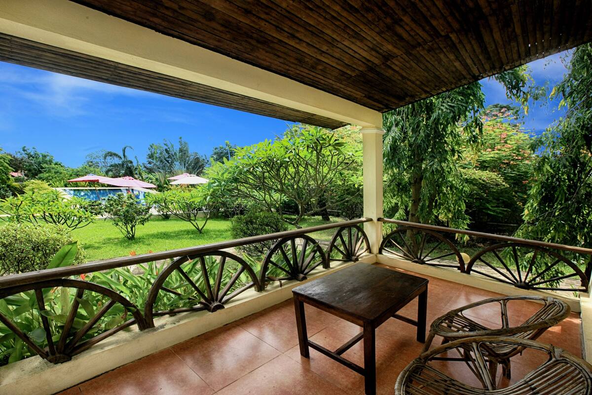 A covered balcony with a view of a lush garden and a building with a red roof.