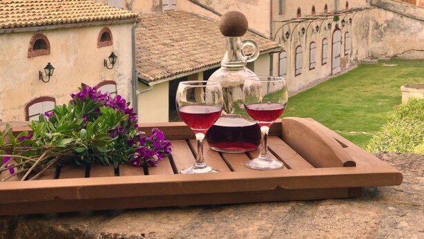 Une terrasse avec une table dressée, agrémentée de verres à vin et d’une carafe, entourée d’un jardin et de bâtiments historiques.
