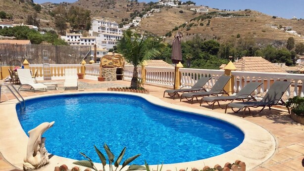 A swimming pool with lounge chairs, a decorative dolphin sculpture, and a view of a hillside with buildings. - CHALET WITH PISICINA AND VIEWS OF DREAM (Almuñécar)
