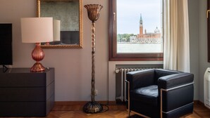 Een moderne kamer met een zwarte leren bank, een vloerlamp, een televisie op een kast, een inlijsting spiegel en een raam met uitzicht op een historisch gebouw.