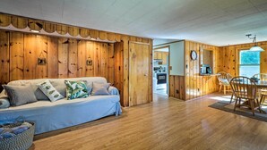 Interior acogedor de una cabaña con pared de madera, un sofá con cojines, una mesa de comedor con sillas y una zona de cocina.