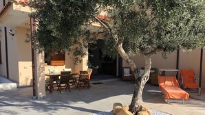 Un patio con una mesa y sillas de comedor, una silla lounge techada y un árbol rodeado de rocas.
