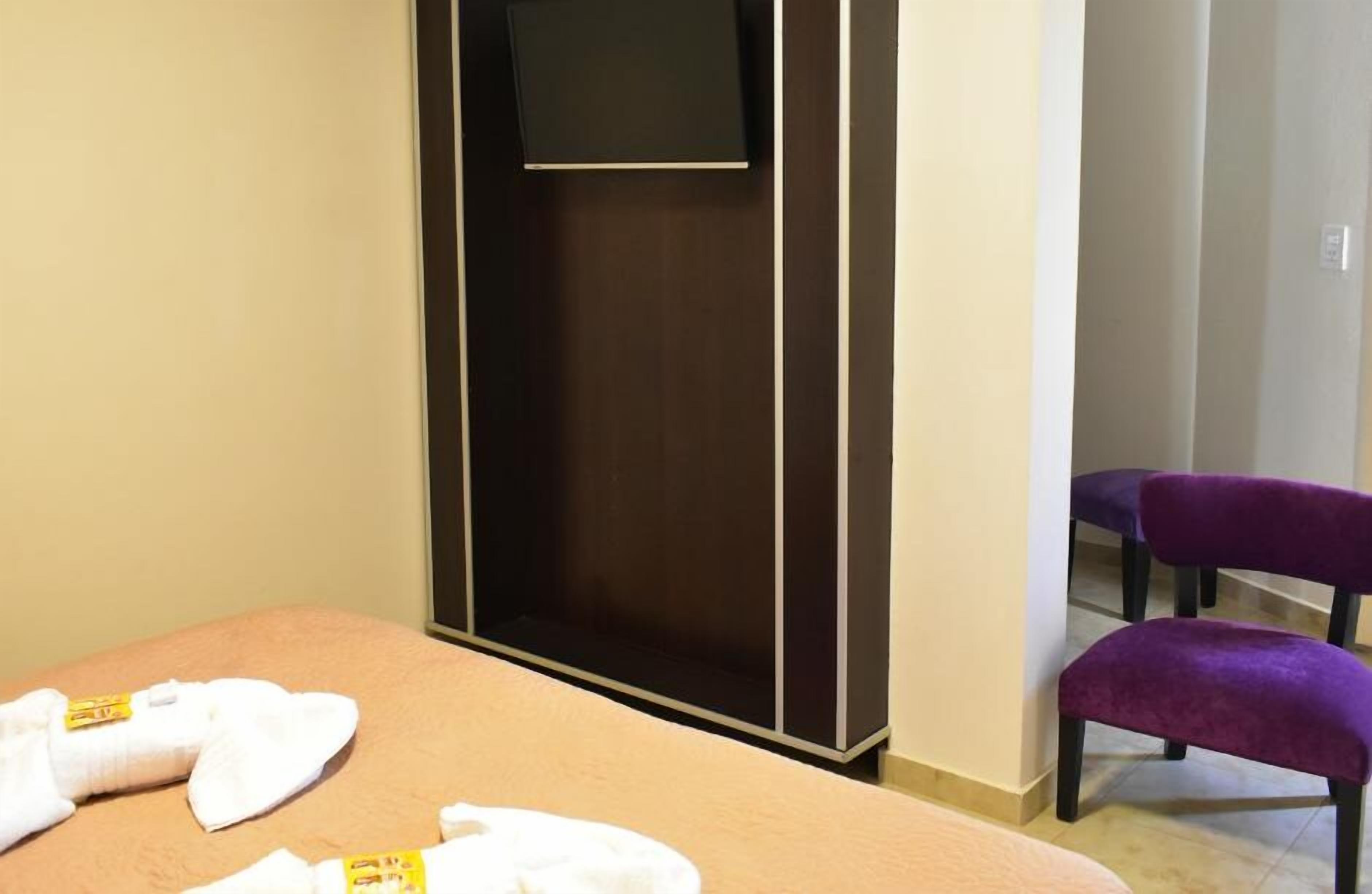 A hotel room with a bed, a TV mounted on a dark wooden cabinet, and two purple chairs.