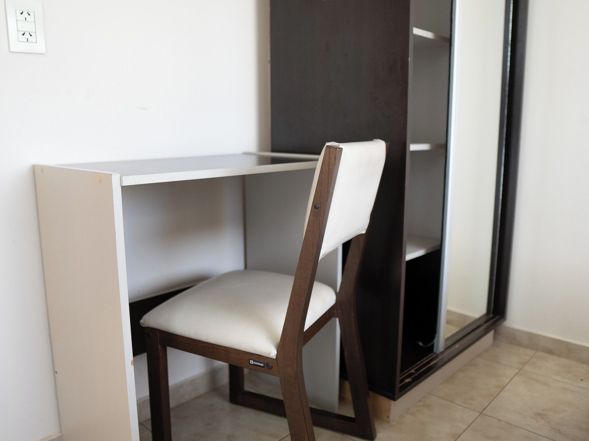 A chair with a wooden frame and white cushion, positioned in front of a white desk with a black shelf.