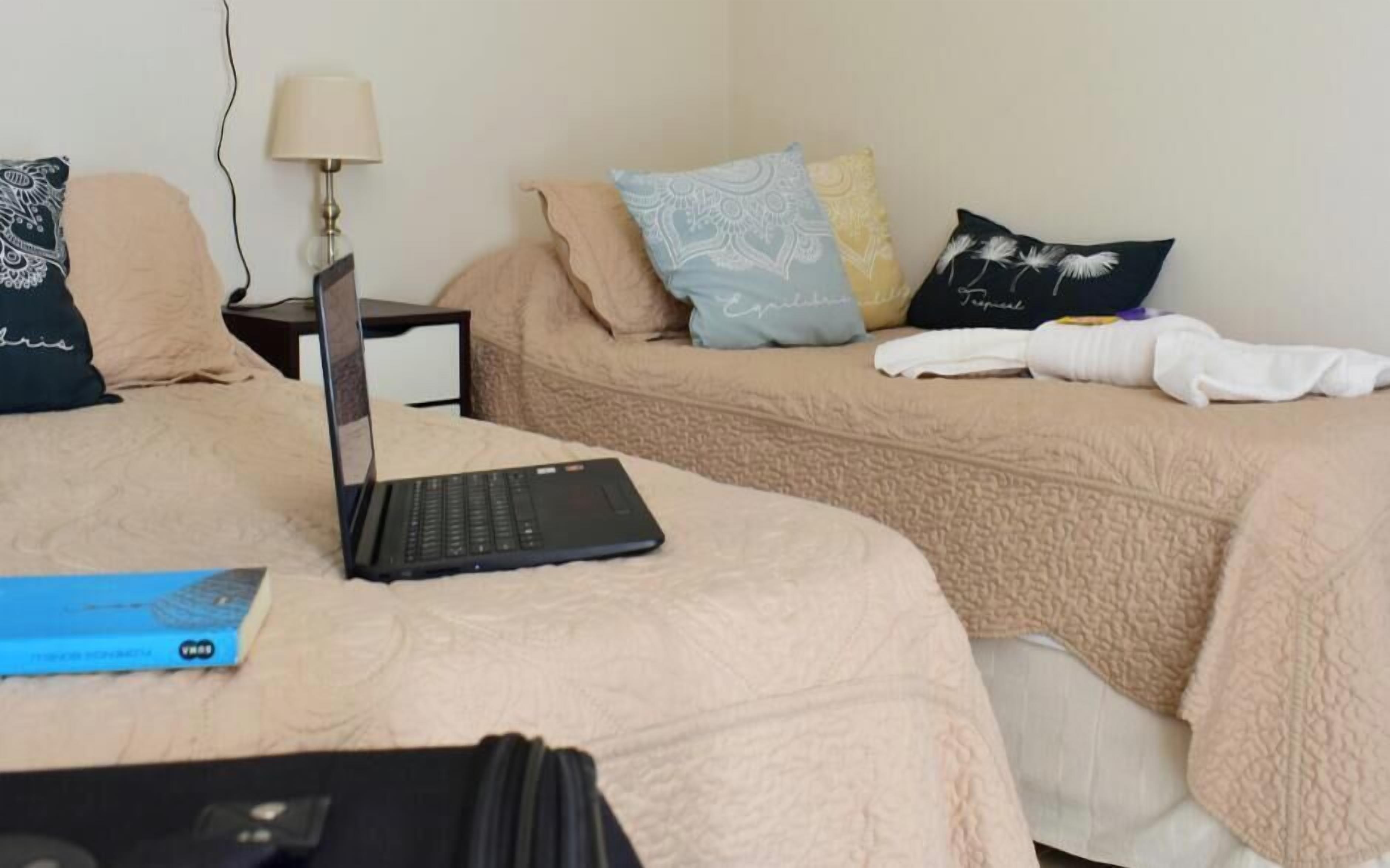 A hotel room with two beds, a laptop on the left bed, a lamp on the nightstand, and a book on the floor.