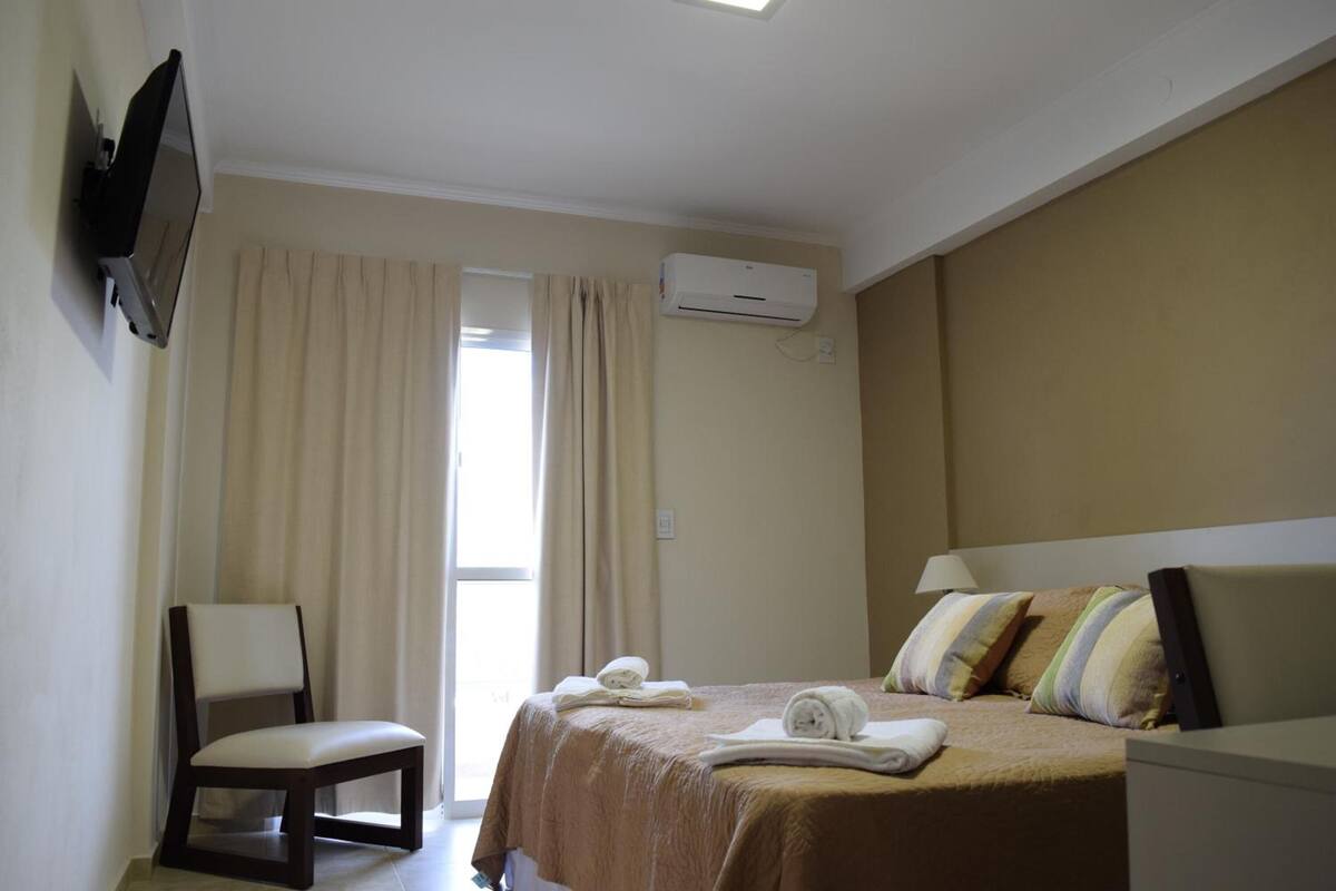 A hotel room with a bed, a chair, a TV mounted on the wall, and an air conditioning unit.