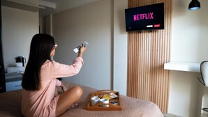 A person sitting on a bed in a hotel room, holding a remote control, with a tray of breakfast items in front of them. A television displaying the Netflix logo is mounted on the wall. - Hotel Rio (Chajarí)