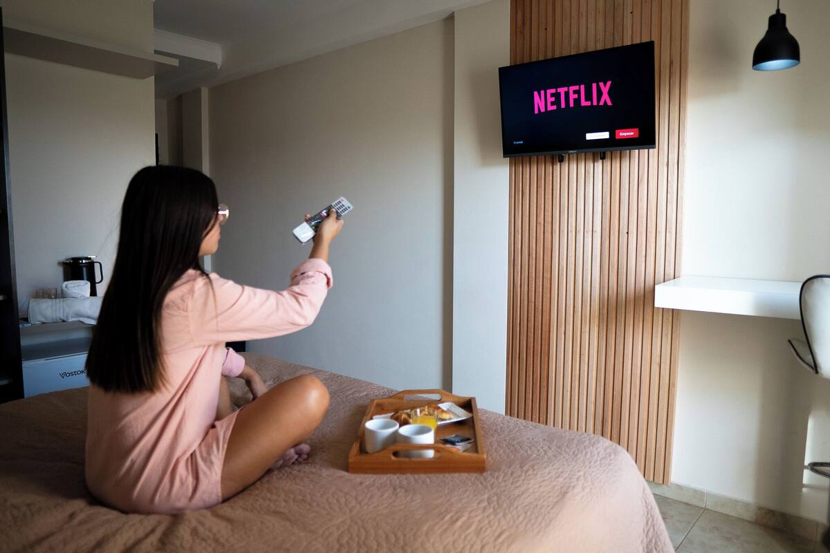 A person sitting on a bed in a hotel room, holding a remote control, with a tray of breakfast items in front of them. A television displaying the Netflix logo is mounted on the wall.