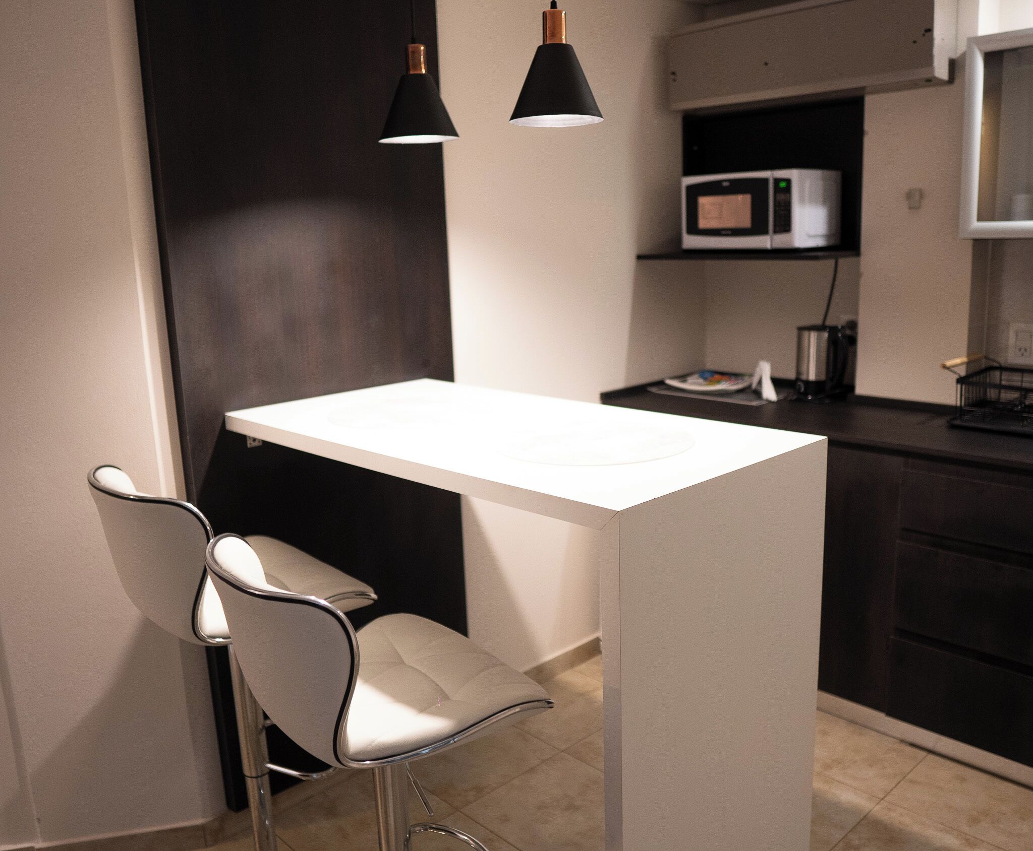 Cozinha moderna com bancada branca, duas luminárias pendentes pretas, micro-ondas e pia de aço inoxidável.