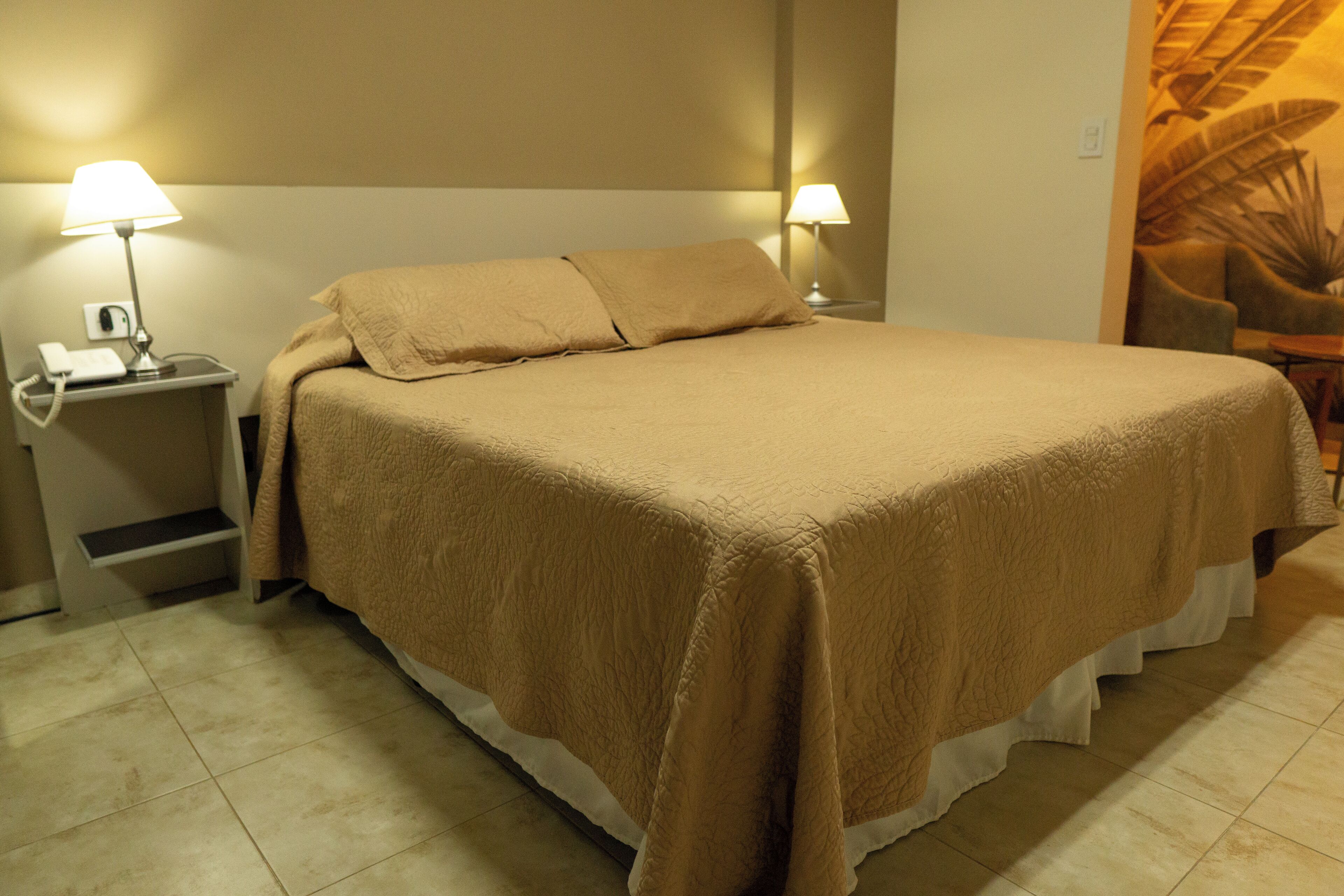 A neatly made bed with a beige blanket and pillows, a bedside lamp, and a nightstand with a phone.