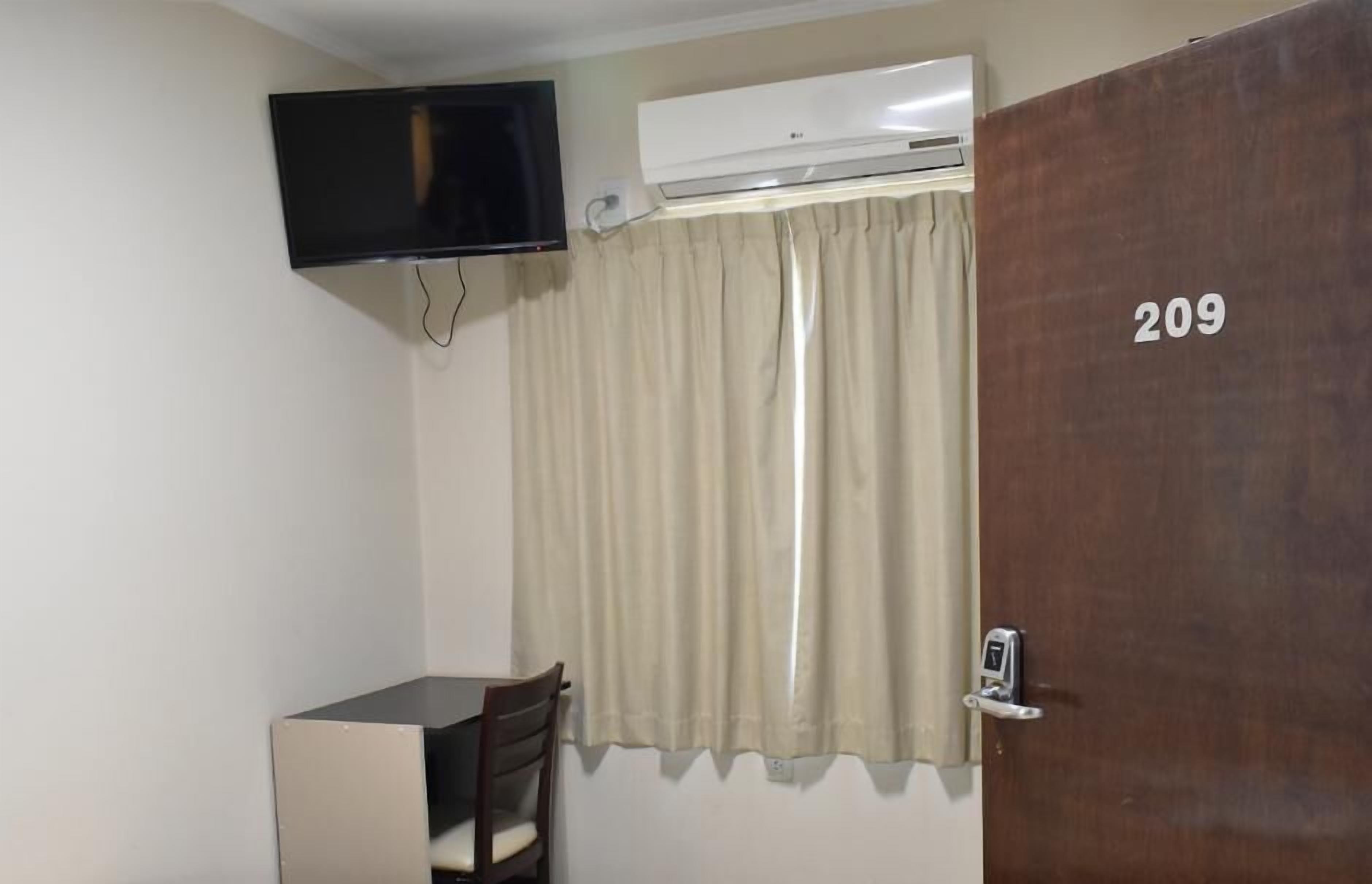 A room with a TV mounted on the wall, an air conditioning unit, a door with the number 209, and a small desk with a chair.