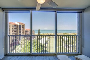 A room with a large sliding glass door leading to a balcony with a view of a beach and buildings.