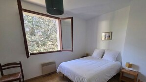 Ein Schlafzimmer mit Bett, Stuhl, Nachttisch, Lampe, Heizkörper und einem Fenster mit Blick auf Bäume.
