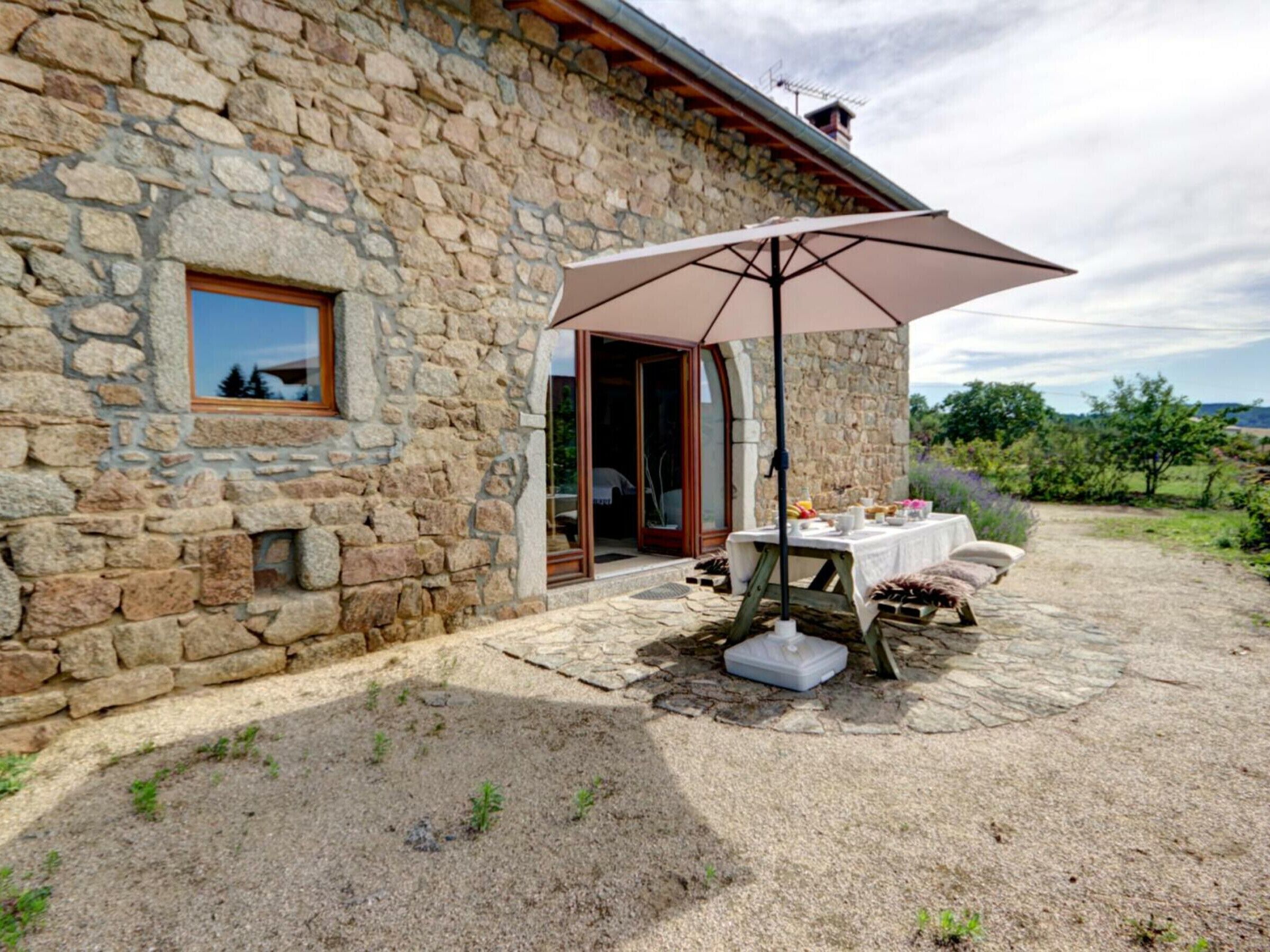 A stone building with a patio area featuring a table set for a meal, an umbrella, and a view of the surrounding countryside.