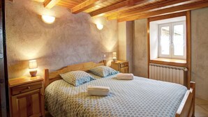 A bedroom with a wooden ceiling, a large bed with patterned bedding, two bedside lamps, and a window with blinds. - Cozy cottage in natural park with pets allowed and modern comfort (Saint-Bonnet-le-Courreau)