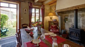 A rustic dining room with a wooden table set for a meal, a stone fireplace, and a view of the garden through the window. - Charming Cottage with Garden in Estivareilles (Estivareilles)